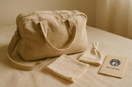 Gepackte Kliniktasche aus Stoff auf hellem Bett mit Baby-Mützchen, Mulltuch und Mutterpass – ruhige Vorbereitung für Geburt im Krankenhaus