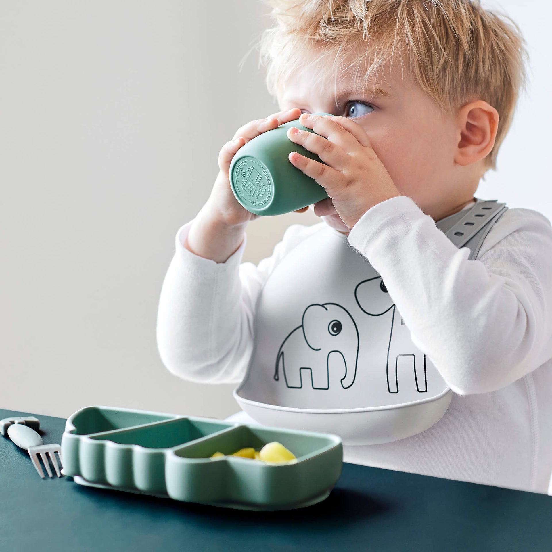 Kleinkind in weißem Pullover mit Elefantenmotiv trinkt aus grünem Becher, vor ihm steht ein grüner, unterteilter Silikonteller mit Babynahrung – BabyLions Ernährung & Unterwegs.