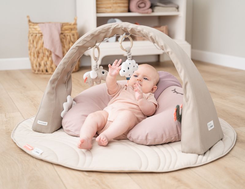Baby liegt entspannt auf einer weichen Spieldecke unter einem beigen Spielbogen mit hängendem Spielzeug aus der Babylions Kollektion.