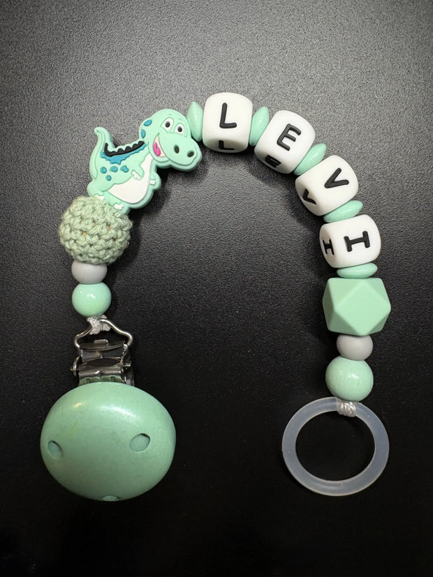 Detailansicht der personalisierten Schnullerkette Dino mit Namen von BabyLions® mit Dino-Motiv, Holzclip, Silikonring sowie Silikon- und Holzperlen in Mint
