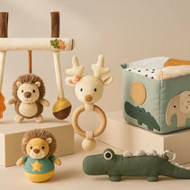 Baby-Spielzeug-Kollektion: Igelanhänger, Reh-Rassel-Greifring, Stoff-Activity-Würfel, Stehauf-Löwe, Krokodil-Kissen auf Podesten.