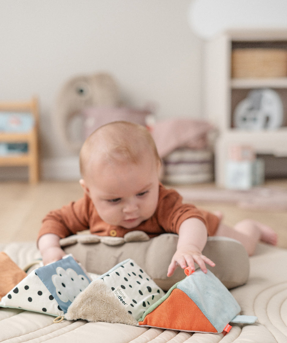 Baby liegt auf dem Bauch und spielt mit einem farbenfrohen, ausklappbaren Stoffwürfel „Deer Friends“ von BabyLions – weiche Stoffelemente in Orange, Blau, Beige und Schwarz-Weiß, ideal zur sensorischen Förderung.
