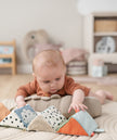 Baby liegt auf dem Bauch und spielt mit einem farbenfrohen, ausklappbaren Stoffwürfel „Deer Friends“ von BabyLions – weiche Stoffelemente in Orange, Blau, Beige und Schwarz-Weiß, ideal zur sensorischen Förderung.