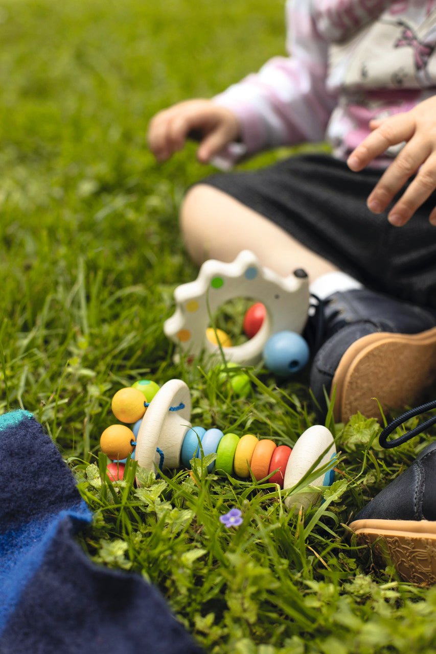 Baby spielt im Gras mit Greifling Elastik Stielrassel Regenbogen aus Holz
